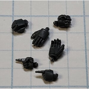 May include: A collection of small, dark gray plastic model hands in various poses. The hands are detailed with sculpted lines and textures, including fists, open palms, and pointing fingers. These are likely miniature accessories for a model kit or action figure.