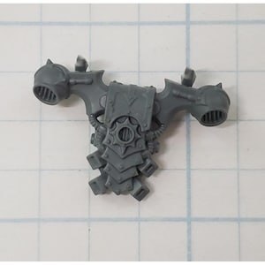 May include: A gray plastic miniature model component, possibly for a tabletop game. It features a central shield-like element with a starburst design, flanked by two rounded, vented structures. The component has intricate details and a textured surface.
