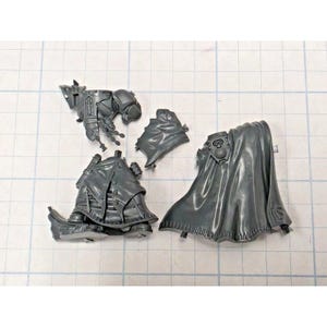 May include: A set of unpainted gray plastic miniature model components including a torso piece with mechanical details, a separate flowing cloak, a lower coat section with button and strap details, and a small shoulder armor plate piece.