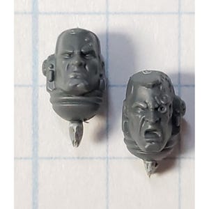 May include: Two grey plastic miniature heads with detailed facial expressions. The heads are for use in a tabletop wargame.