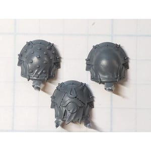 May include: Three gray plastic shoulder pads for a miniature figure. One shoulder pad has a design of a circle with a flame inside. The other two shoulder pads have a design of a circle with many small dots.