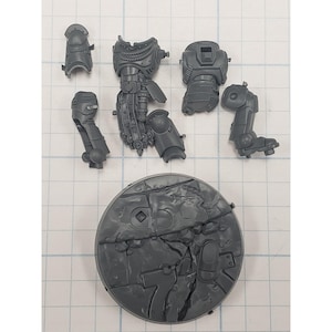 May include: A collection of unpainted, gray plastic miniature parts for a tabletop game. The components include a round base, torso, arm, leg, and shoulder armor pieces. The parts are detailed with mechanical and armored designs.