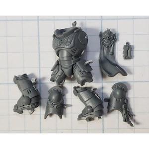 May include: A collection of unpainted, gray plastic miniature armor pieces, including a torso, leg armor, shoulder pads, and a cape. The pieces feature intricate details, such as sculpted wings, skulls, and decorative trim, suggesting a fantasy or sci-fi theme.