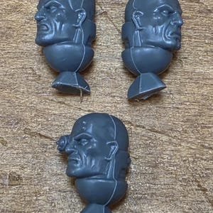 May include: Three unpainted gray plastic miniature heads for gaming figures. Two heads feature distinct facial expressions and hairstyles, while the third head includes a circular ocular implant over one eye. Each piece has a neck joint base.