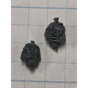 May include: Two unpainted dark gray plastic miniature heads. One head features a detailed mechanical helmet with a respirator and a small top fin. The other head is a bare, bald human-like character with a stern expression and a small top knot.