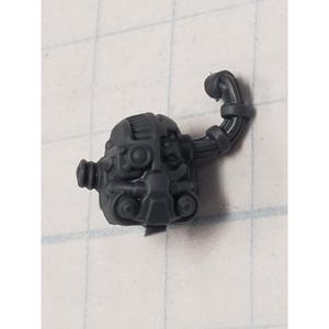 May include: A dark gray plastic miniature component, possibly for a model kit. The piece features a complex, mechanical design with various cylindrical and rounded elements, and a curved, ribbed tube extending from the main body.