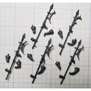 May include: Several dark gray miniature weapons and armor pieces are arranged on a grid background. The weapons include spears with curved blades and ornate detailing. Additional components include gauntlets and helmet-like pieces.