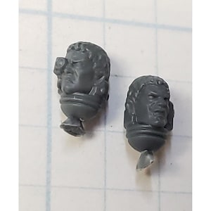 May include: Two gray plastic miniature heads with detailed facial features. One head has a small, rectangular object attached to the side of the head.
