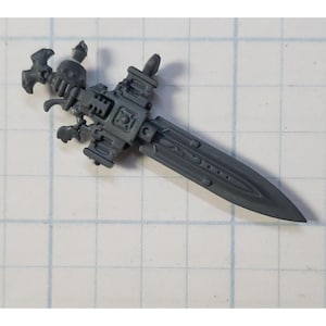 May include: A gray plastic model of a futuristic sword with a detailed design. The sword has a handle with a skull-like design and a blade with intricate carvings.