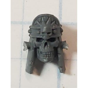 May include: A gray plastic skull with a decorative headband. The skull has a toothy grin and a detailed design on the headband.