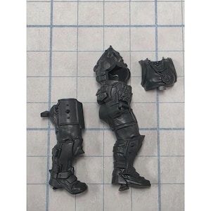 May include: Three unpainted dark gray plastic miniature model components consisting of a separate leg piece, a torso piece with integrated head and backpack, and a chest plate piece. These parts feature detailed armor plating and mechanical textures.