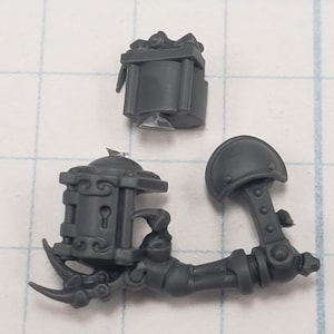 May include: Three-piece, dark gray miniature model kit. Includes a rectangular box, a mechanical arm with a claw, and a shoulder pad. The arm features a detailed, ornate design with a keyhole and decorative elements. Ideal for hobbyists and model builders.