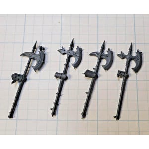 May include: Four dark gray plastic miniature polearm weapons, each featuring a central axe blade, a pointed top spike, and a textured handle grip. These unpainted gaming components display intricate molded details including small studs and decorative elements.