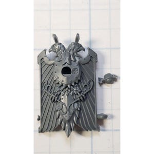 May include: A gray plastic model of a double-headed eagle shield with a circular hole in the center. The shield is detailed with feathers, leaves, and a decorative design. The model is for a miniature wargame.