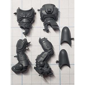 May include: A collection of unpainted, gray plastic miniature model parts. The components include armored torso pieces, leg sections, and knee guards, likely for a tabletop war game. The parts are detailed with mechanical and decorative elements.