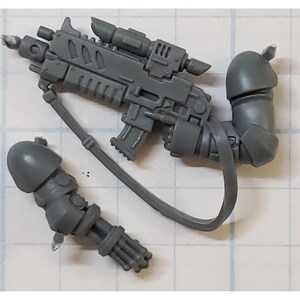 May include: A collection of gray plastic miniature weapon components, including a rifle with a scope, a shoulder-mounted arm, and a multi-barreled gun arm. The pieces are detailed and appear to be from a model kit.