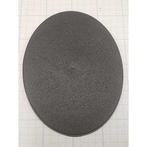 May include: A dark gray, oval-shaped object with a textured surface. The object has a slightly raised central point. The item is set against a white grid background.