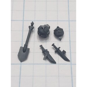 May include: A collection of gray plastic miniature accessories, including a shovel, two swords, and two spherical objects. The items are arranged on a grid-lined surface, likely for scale or organization.