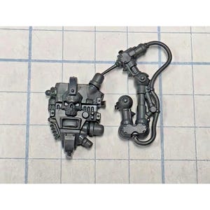 May include: A dark gray plastic miniature backpack component featuring a prominent skull emblem on the central housing, connected by a curved cable to a mechanical arm assembly with multiple joints and a circular attachment point at the base.