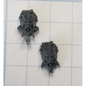 May include: Two gray plastic miniature heads with detailed facial features and a textured surface. The heads have a menacing expression with sharp teeth and a pointed chin.