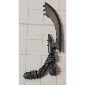 May include: A dark gray miniature weapon featuring a curved blade with three triangular teeth. The weapon is held by a skeletal hand and arm, with a cylindrical grip at the base. The overall design suggests a fantasy or tabletop game theme.
