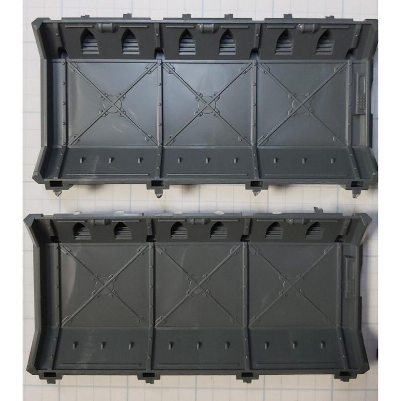 May include: Two gray plastic wall sections with a grid pattern and small rectangular openings. The sections are designed for use in a model building or miniature gaming set.