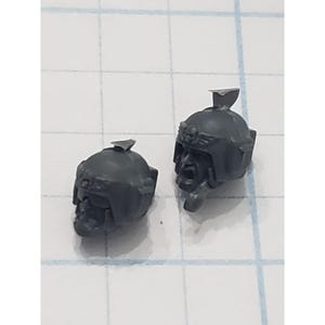 May include: Two gray plastic miniature helmets with detailed facial features and winged emblems. Each helmet has a small, angled visor. The helmets are likely for tabletop gaming or model building.