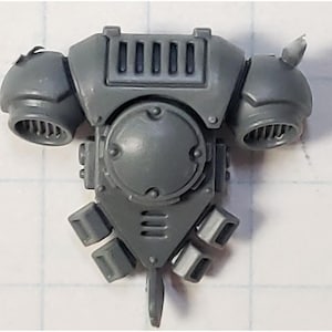 May include: A gray plastic miniature model of a futuristic armored torso with shoulder pads and exhaust vents. The model has a circular chest plate and rectangular details, likely for a tabletop game or hobby project.