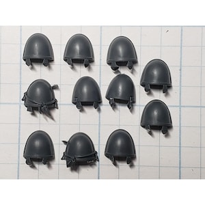 May include: A collection of ten dark gray plastic shoulder pads, each with a rounded dome shape and attached lower components. Some pads have additional details. The items are arranged on a grid-lined surface, suggesting a crafting or hobby context.