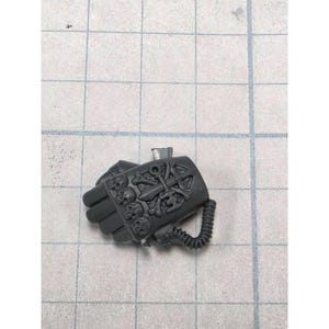 May include: A dark gray miniature power fist accessory featuring a sculpted design of three skulls and a central sword emblem with crossed bones. The piece includes a flexible ribbed cable attachment on the side, crafted from plastic material.