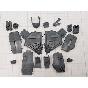 May include: A collection of gray plastic model kit parts, including large armor panels, smaller components, and various mechanical elements. The pieces are arranged on a white grid surface, suggesting a construction or assembly theme.