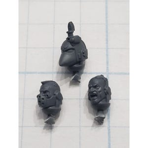 May include: Three gray plastic miniature heads, likely for a tabletop game. One head is helmeted with a pointed visor and antenna. Another has a muzzle-like mask, and the third is a screaming face.