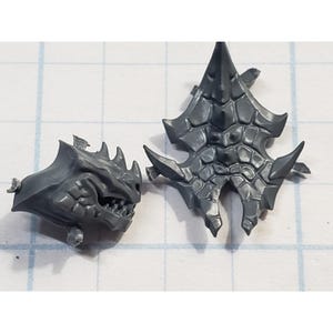 May include: Two gray plastic miniature fantasy game components. One is a horned head with a snarling mouth, the other is a spiky shield-like piece. Both have a textured, armored appearance, suggesting a creature or character.