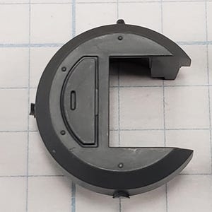 Puede incluir: Un componente de pl&aacute;stico gris oscuro con una forma exterior circular y una secci&oacute;n rectangular. El componente tiene un peque&ntilde;o detalle en relieve y varias peque&ntilde;as hendiduras circulares. El fondo es un patr&oacute;n de cuadr&iacute;cula.