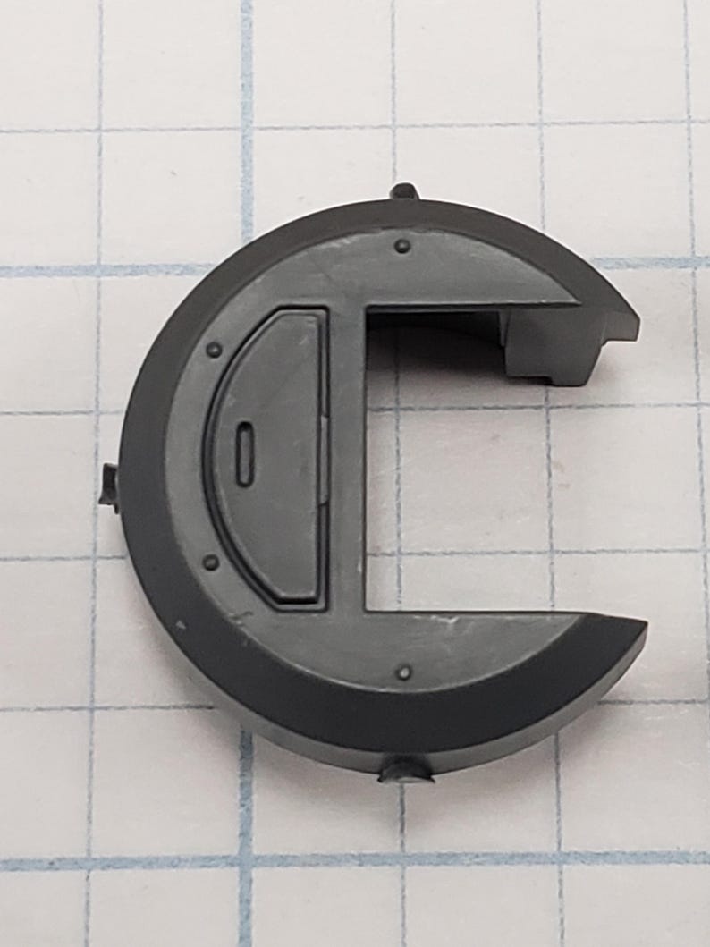 Puede incluir: Un componente de pl&aacute;stico gris oscuro con una forma exterior circular y una secci&oacute;n rectangular. El componente tiene un peque&ntilde;o detalle en relieve y varias peque&ntilde;as hendiduras circulares. El fondo es un patr&oacute;n de cuadr&iacute;cula.