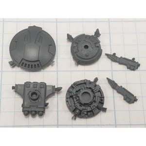 May include: A collection of six unpainted gray plastic model kit components, including a large circular shield, a rectangular engine block with three vents, two smaller circular mechanical parts, and two elongated weapon pieces with pointed tips.