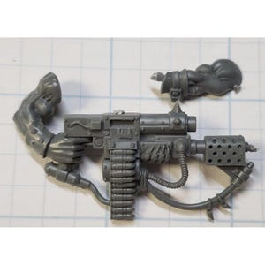 May include: A gray plastic model of a futuristic weapon with a belt of ammunition, a scope, and a handle. The weapon has a detailed design with a variety of textures and shapes.