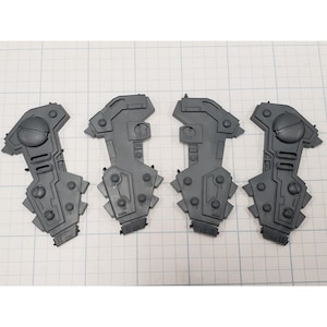 May include: Four gray plastic model components, possibly for a larger structure or vehicle. Each piece has a unique shape with a combination of flat and angled surfaces, along with circular and rectangular details. The components are arranged on a grid.