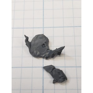 May include: Two gray plastic model pieces, possibly from a miniature figure or vehicle. One piece has a curved, organic shape with a square indentation. The other piece is smaller and angular.