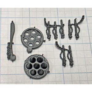 May include: A collection of dark gray plastic model kit components including a long curved piece with ornate detailing, two circular plates with six-hole patterns, and three separate sections of vertical bars connected by twisted rope-like structures.