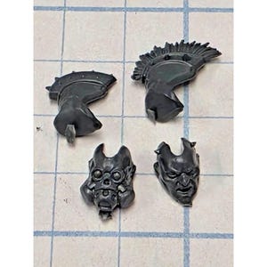 May include: Four unpainted dark gray plastic miniature components consisting of two helmet crests with spiked edges and two distinct head pieces, one featuring a skull-like mask with circular eye sockets and the other a human-like face with a grim expression.