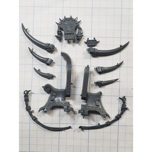 May include: A collection of gray plastic miniature model parts, including horns, decorative armor, and curved chain-like pieces. The components are arranged on a grid-lined surface, suggesting a hobby or model-making theme.