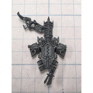 May include: A dark gray plastic miniature component featuring a central vertical tower structure, two skull-like side ornaments, and a flowing smoke-like plume extending from the top. The piece includes intricate molded details and sharp edges.