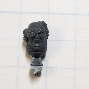 May include: A gray plastic miniature head with a single eye and a distressed face. The head is attached to a small gray plastic base.