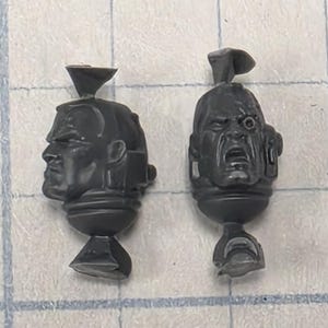 May include: Two unpainted dark gray plastic miniature heads. The left head is in profile, while the right head faces forward with an open mouth and a mechanical eyepiece. Both pieces feature a small sprue attachment point at the top and bottom.