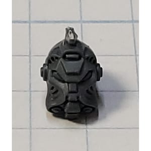 May include: A dark gray plastic miniature helmet with intricate details, including vents, a central visor area, and a raised crest. The helmet appears to be a component of a larger model or figure, likely for a tabletop game or hobby project.