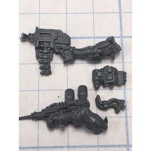 May include: A collection of dark gray plastic miniature model components including a firearm with an attached arm, a separate rectangular mechanical device, a second firearm with a long barrel and two top cylinders, and a small curved exhaust pipe piece.