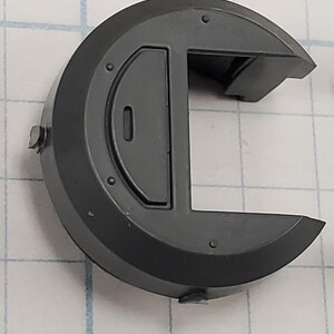 Puede incluir: Un componente de pl&aacute;stico gris oscuro, semicircular, con una incrustaci&oacute;n rectangular y una secci&oacute;n recortada. El componente tiene varios remaches circulares peque&ntilde;os y una peque&ntilde;a protuberancia en la parte superior. El objeto est&aacute; sobre un fondo de cuadr&iacute;cula.