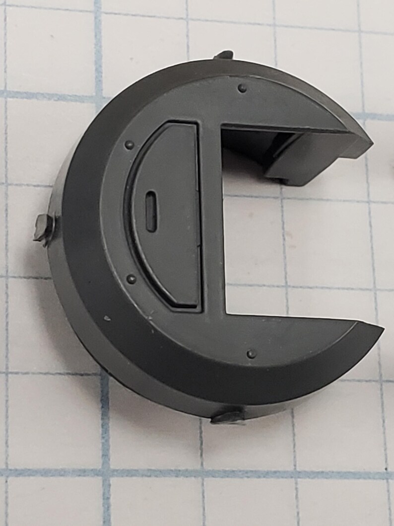 Puede incluir: Un componente de pl&aacute;stico gris oscuro, semicircular, con una incrustaci&oacute;n rectangular y una secci&oacute;n recortada. El componente tiene varios remaches circulares peque&ntilde;os y una peque&ntilde;a protuberancia en la parte superior. El objeto est&aacute; sobre un fondo de cuadr&iacute;cula.