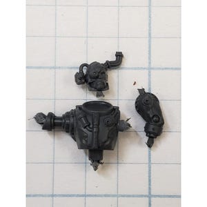 May include: Three dark gray plastic miniature model parts, possibly for a science fiction or fantasy game. The parts include a head, a torso, and an arm, all with intricate mechanical details and a weathered appearance.